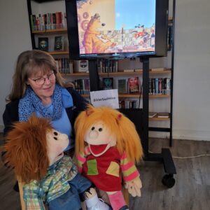 Helga Rebling sitzt mit zwei Handpuppen auf dem Schoß vor einem Monitor, auf dem ein Bild mit einem Bären aus einem Bilderbuch zu sehen ist.