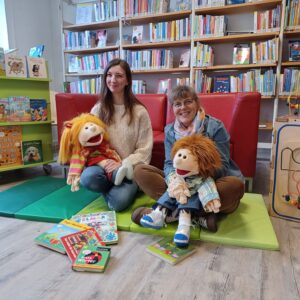 Gina Friedrichs und Helga Rebling sitzen mit den Stoffpuppen Lilabelle und Fabian auf dem Schoß in der Kinderbücherei und lächeln in die Kamera.